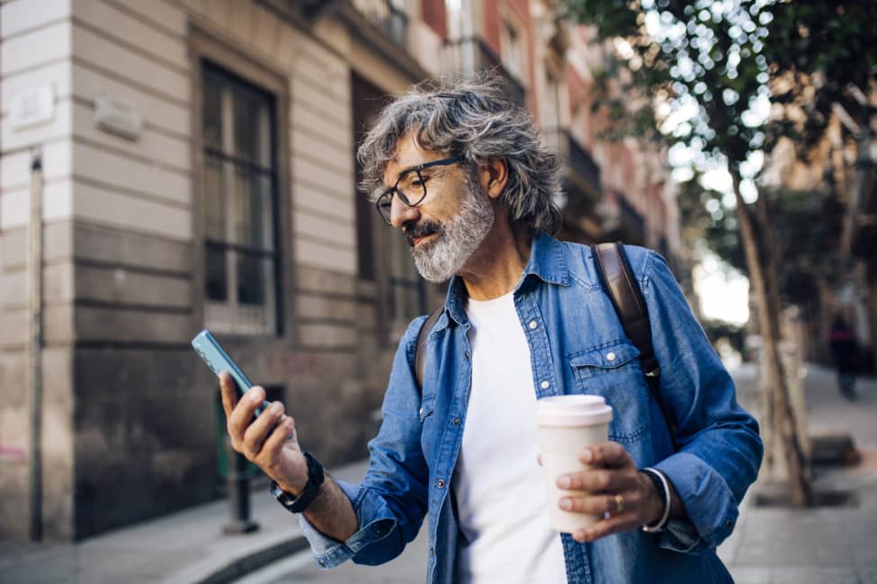 Older man with a coffee and a backpack searching for something on his phone.