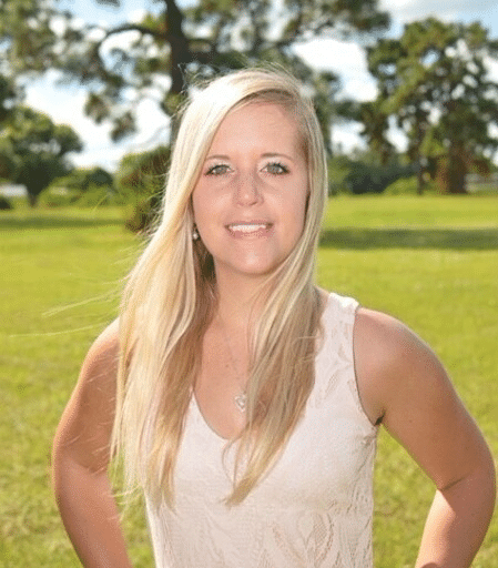 Headshot of a person smiling and looking directly at the camera. They have long blonde hair and are wearing a light-colored shirt. The background is green grass and trees. The person's expression is friendly and approachable.